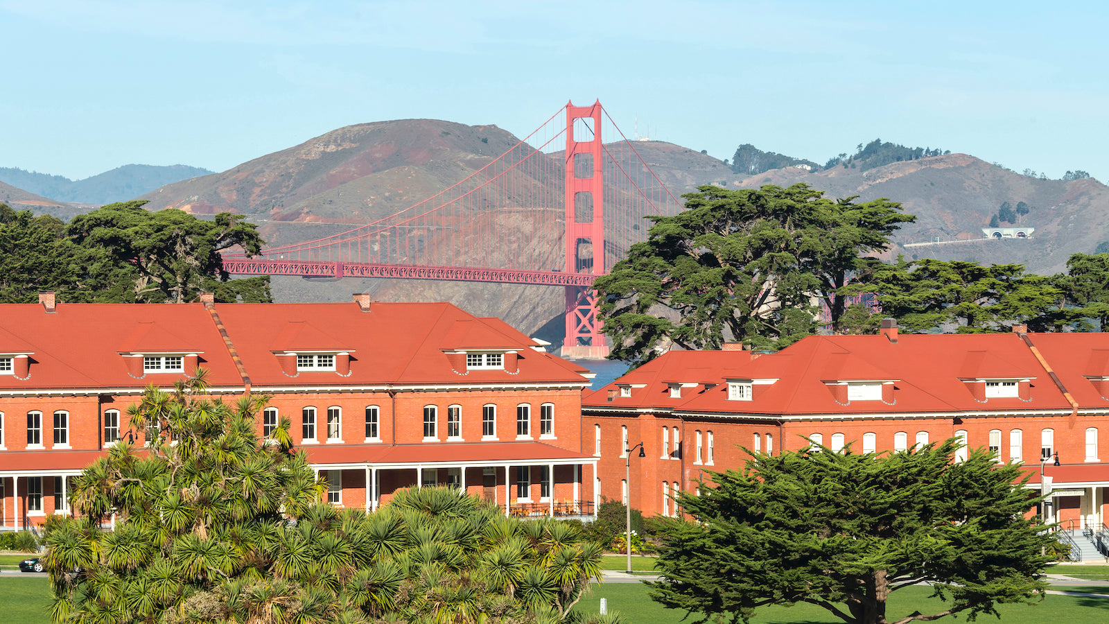 View of the Presidio of San Francisco on a sunny day, looking towards the Golden Gate Bridge. Red brick barracks with white trim are clustered among lush green trees.