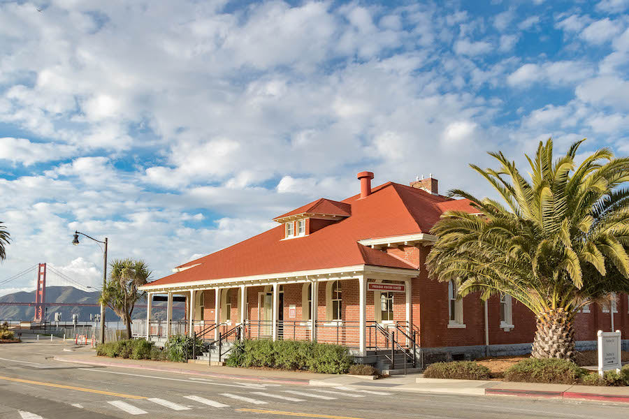 A view of the Presidio Visitor Center on a sunny day, bright green palm trees in the foreground, and a brightly lit Golden Gate Bridge in the background.