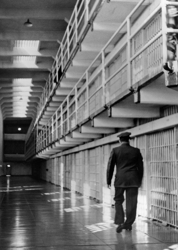 Black and white historical photo of Alcatraz cellblock corridor, with guard walking next to cells