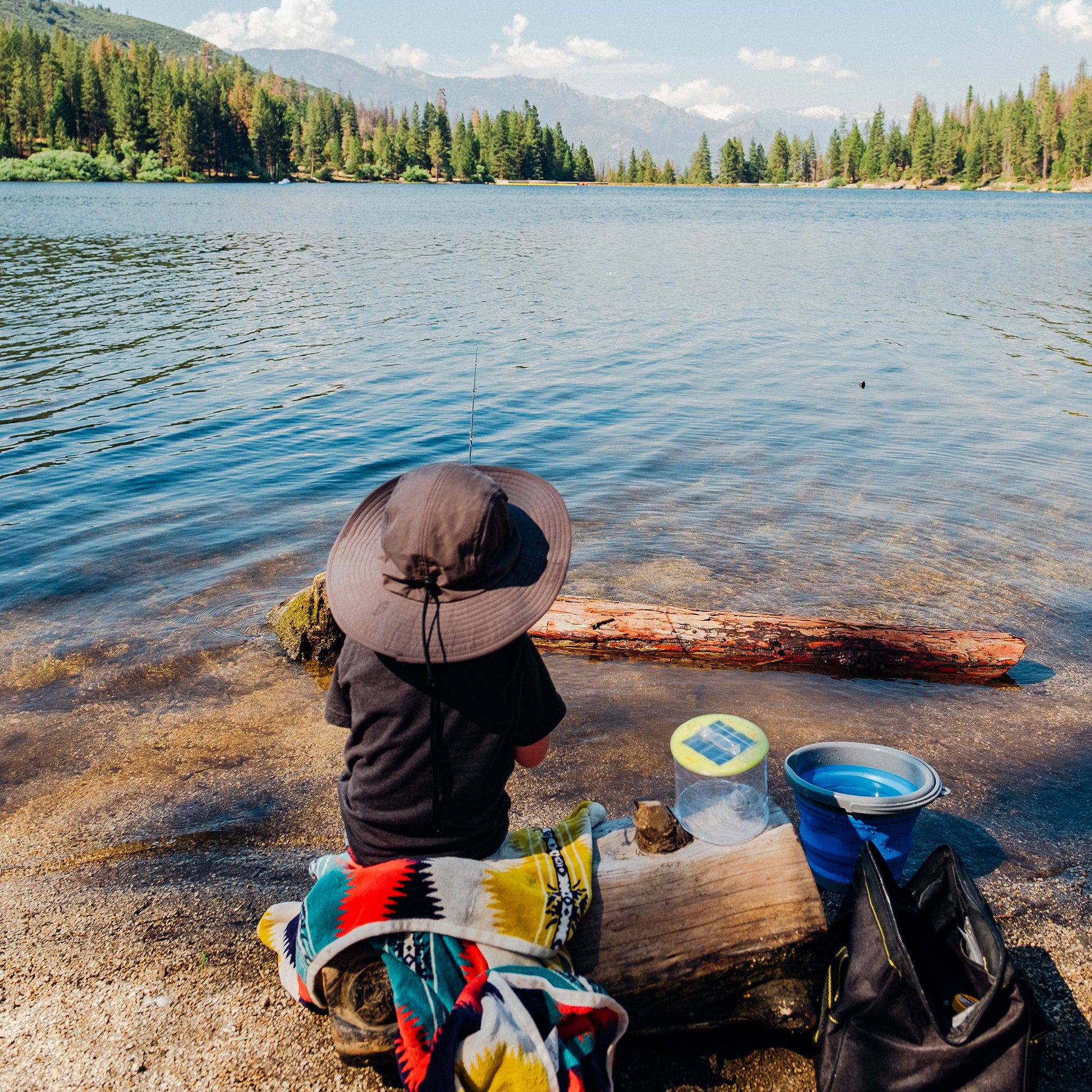 Child sitting on log next to a scenic lake surrounded by mountains and trees. On the log, there are a blanket, bucket, bag, and rechargeable Luci solar light.