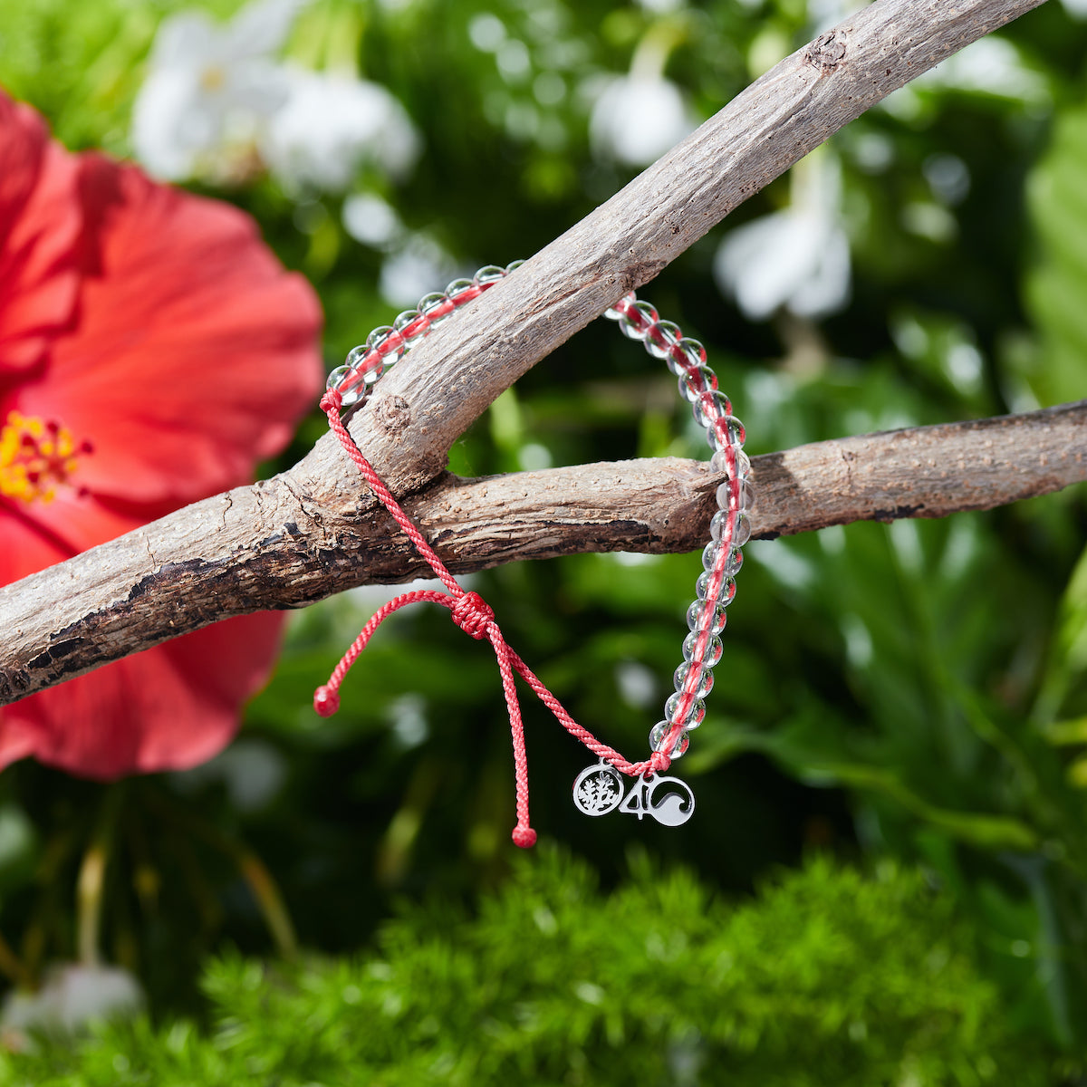 4Ocean Hawaiian Coral Reef Bracelet, with red recycled plastic cord, colorless recycled glass beads, and recycled steel metal charms. It is hanging on a tree branch in front of a bright red hibiscus flower and lush green background.