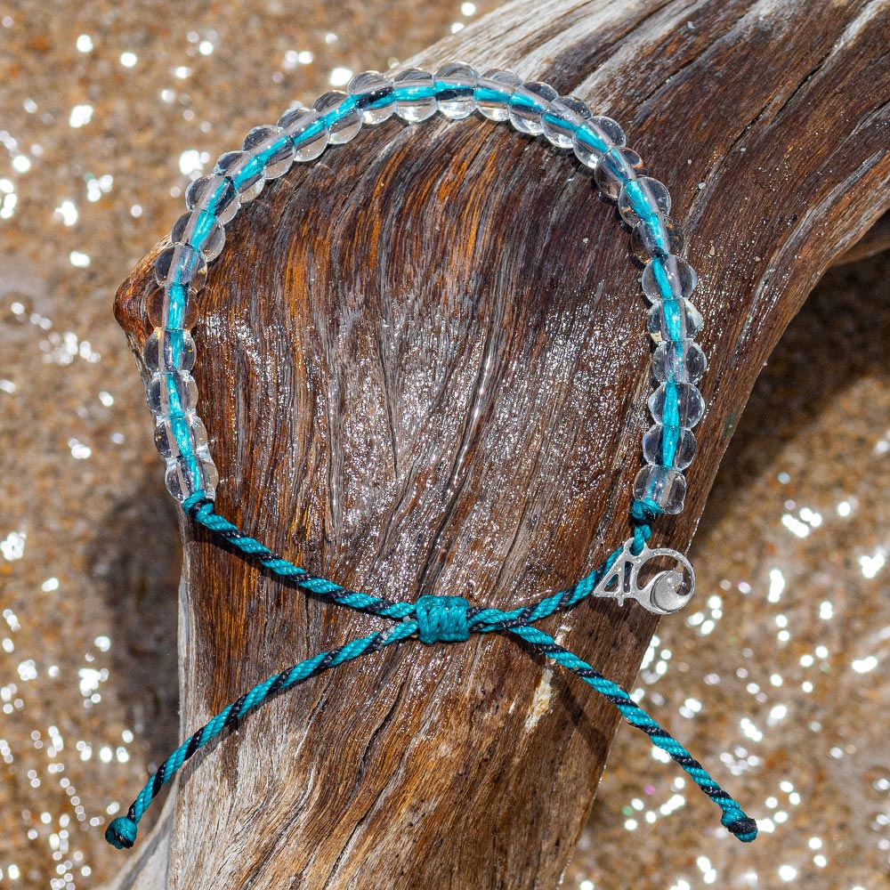 4Ocean Sea Otter Bracelet, with blue and black recycled plastic cord, colorless recycled glass beads, and recycled steel metal charm. It is lying on a wooden log, surrounded by sand and sparkling water.