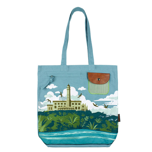 Blue tote bag with an embroidered design of Alcatraz Island, including prison buildings, plants, birds, and fog.