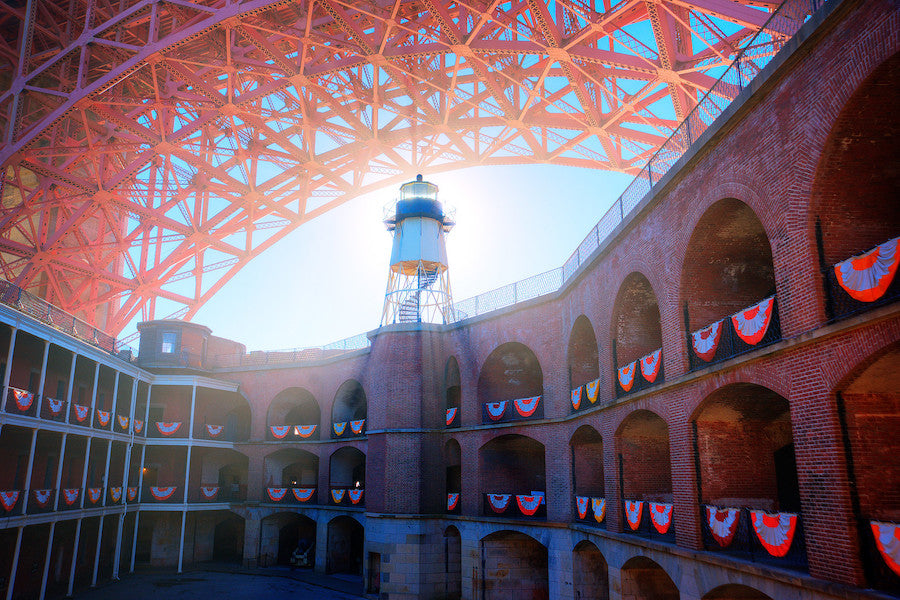 A dramatic look up from within the courtyard of San Francisco’s Fort Point. Patriotic red, white, and blue bunting adorn the brick edifice while the metalwork of the Golden Gate Bridge soars above.