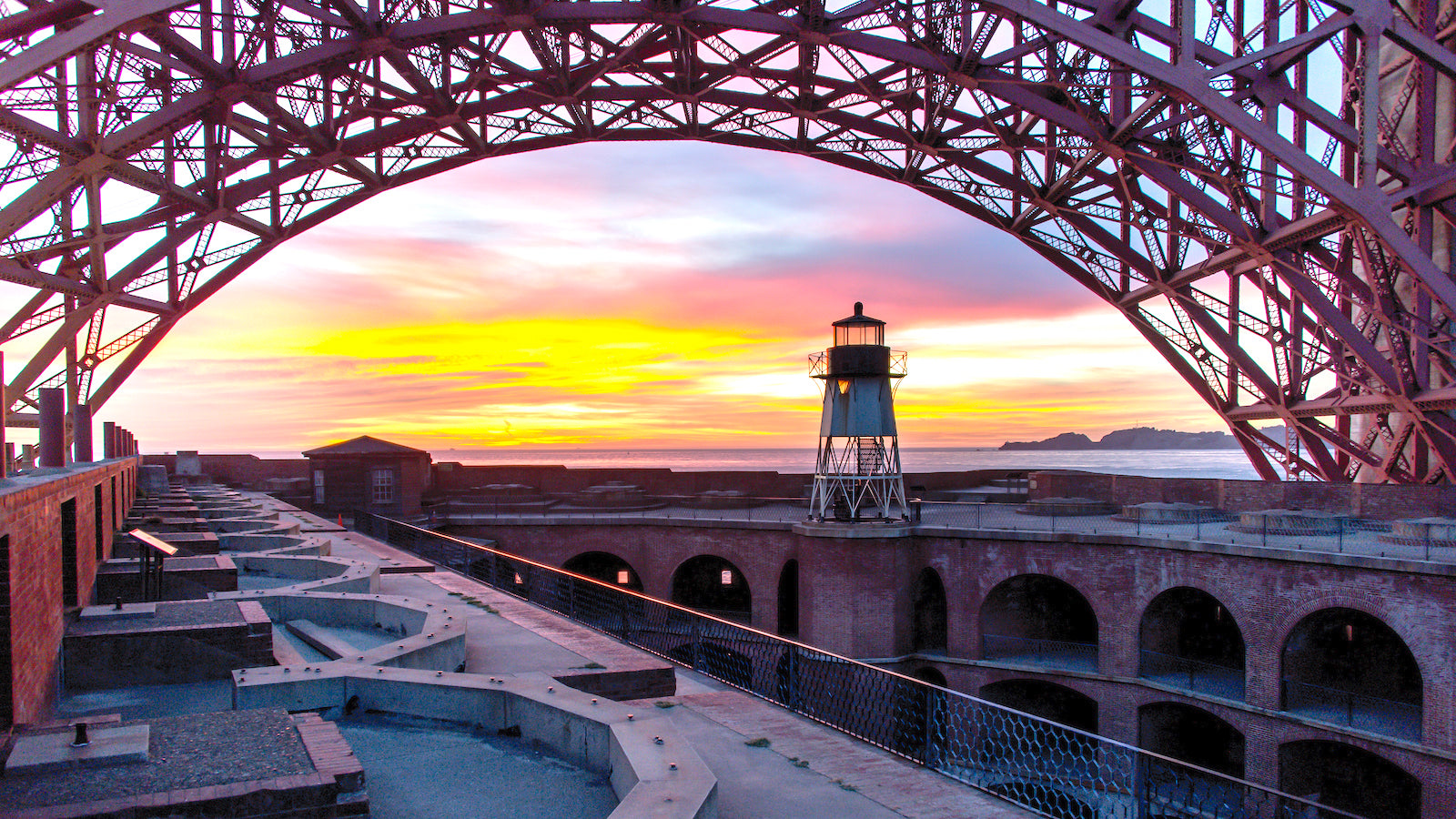 A dramatic view from the roof of Fort Point at sunset. The clouds are brightly lit pink, yellow, and purple, while the steelwork of the Golden Gate Bridge soars above.