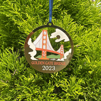 Limited edition 2023 commemorative ornament by the Golden Gate National Parks Conservancy, a circular gold ornament with multicolor Golden Gate Bridge giclee print design, hanging on a tree