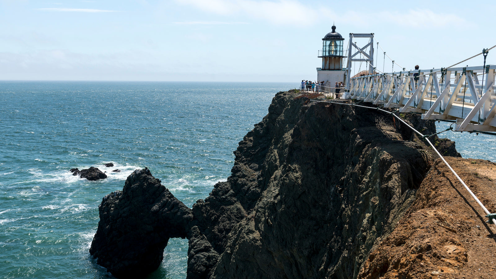 A dramatic view of Point Bonita lighthouse in the Marin Headlands. Rocky cliffs descend into turquoise waters, while the soaring pedestrian bridge rises in the foreground.