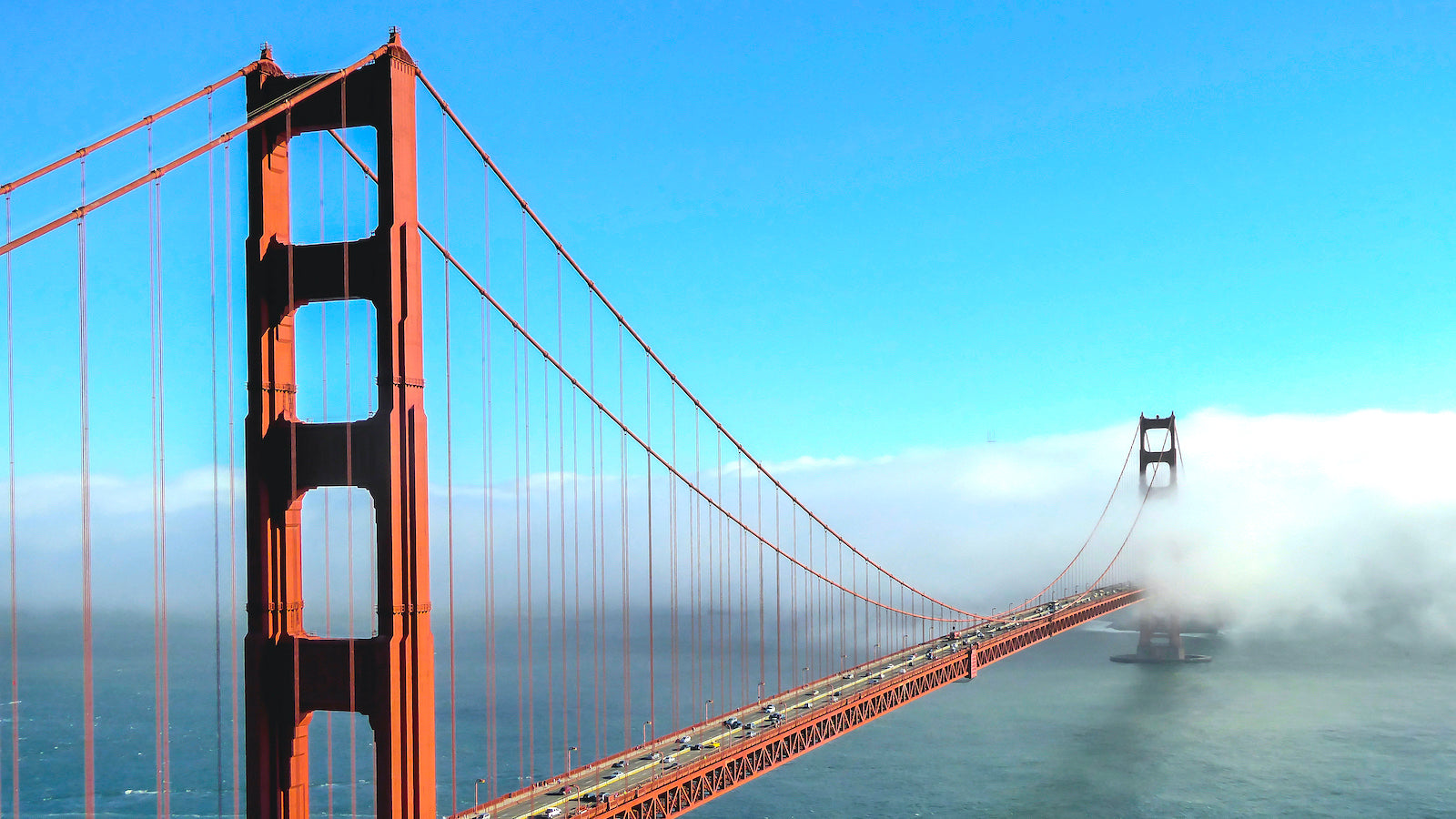 A soaring view of the Golden Gate Bridge on a sunny day, with low fog in the background.