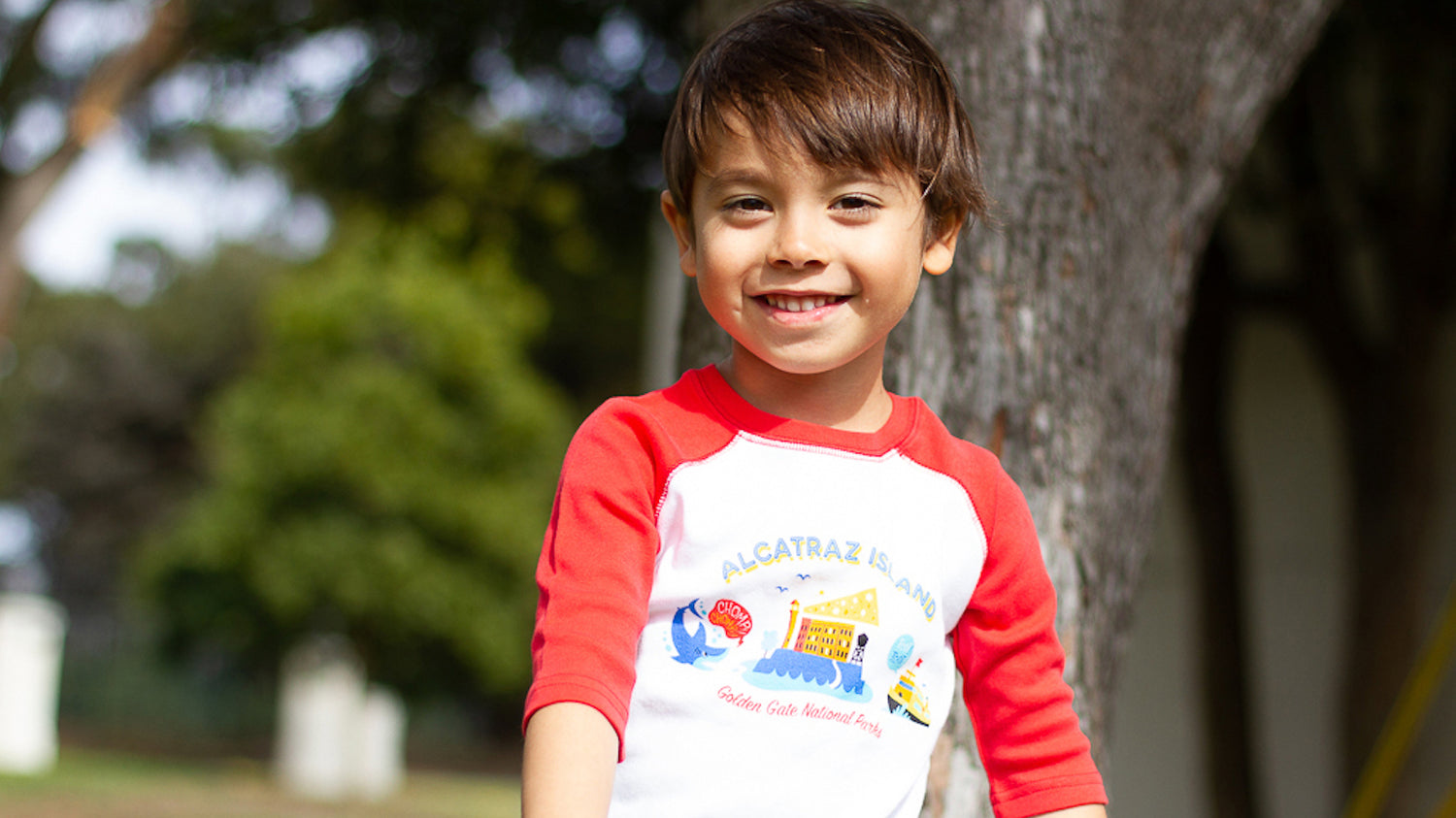 Smiling boy stands in a park looking at the camera. He wears a bright red and white 3/4 raglan sleeve t-shirt, with a colorful Alcatraz Island illustration on the chest.