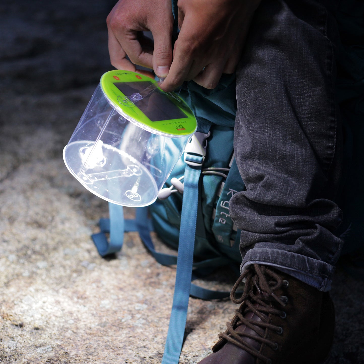 Person holding a rechargeable Luci solar light, lit at night. It is near the ground, illuminating their backpack, jeans, and hiking boots.