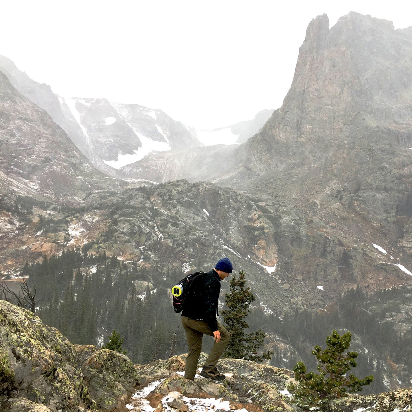 A man hikes in high altitude scenic mountains covered in snow. He wears a beanie and carries a backpack with a rechargeable Luci solar light attached.