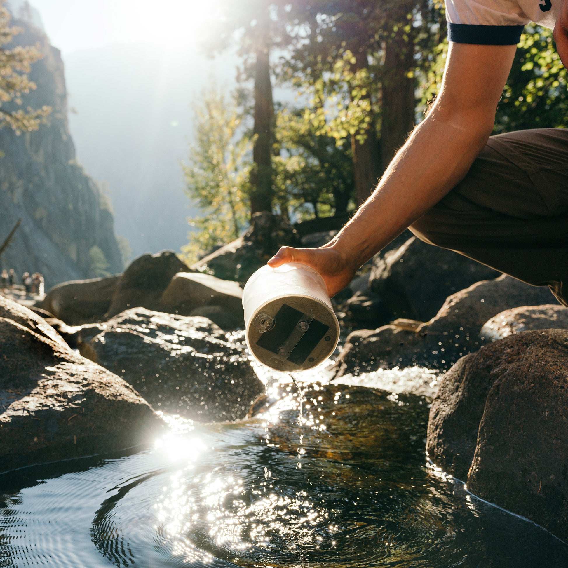 A man washes a rechargeable Luci solar light in a scenic stream, surrounded by misty mountains and lush forest.
