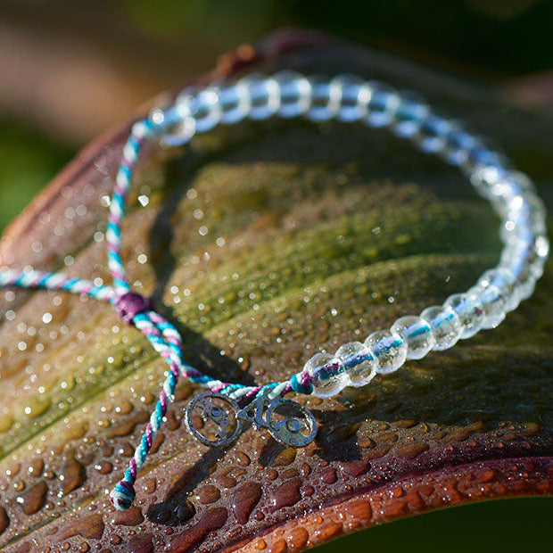 4Ocean Dwarf Sperm Whale Bracelet, with blue, purple, and white recycled plastic cord, colorless recycled glass beads, and recycled steel metal charms. It is lying on a tropical plant leaf, covered in dew.