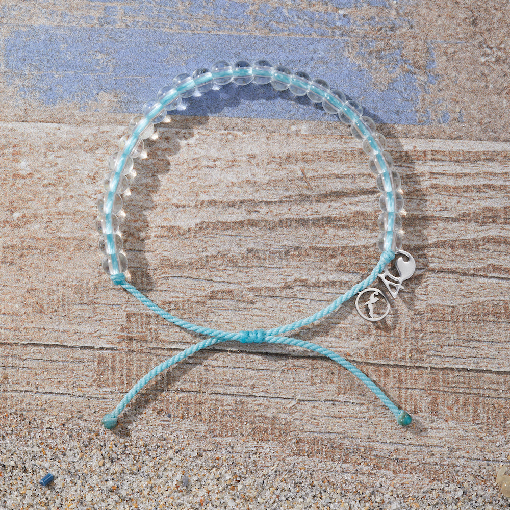 4Ocean Black-Capped Petrel Bracelet, with blue recycled plastic cord, colorless recycled glass beads, and recycled steel metal charm. It is lying on a wooden board background with sand surrounding it.