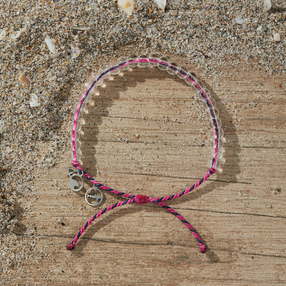4Ocean Blue Shark Bracelet, with pink and black recycled plastic cord, colorless recycled glass beads, and recycled steel metal charms. It is lying on a wooden background, surrounded by sand and small pieces of shell.