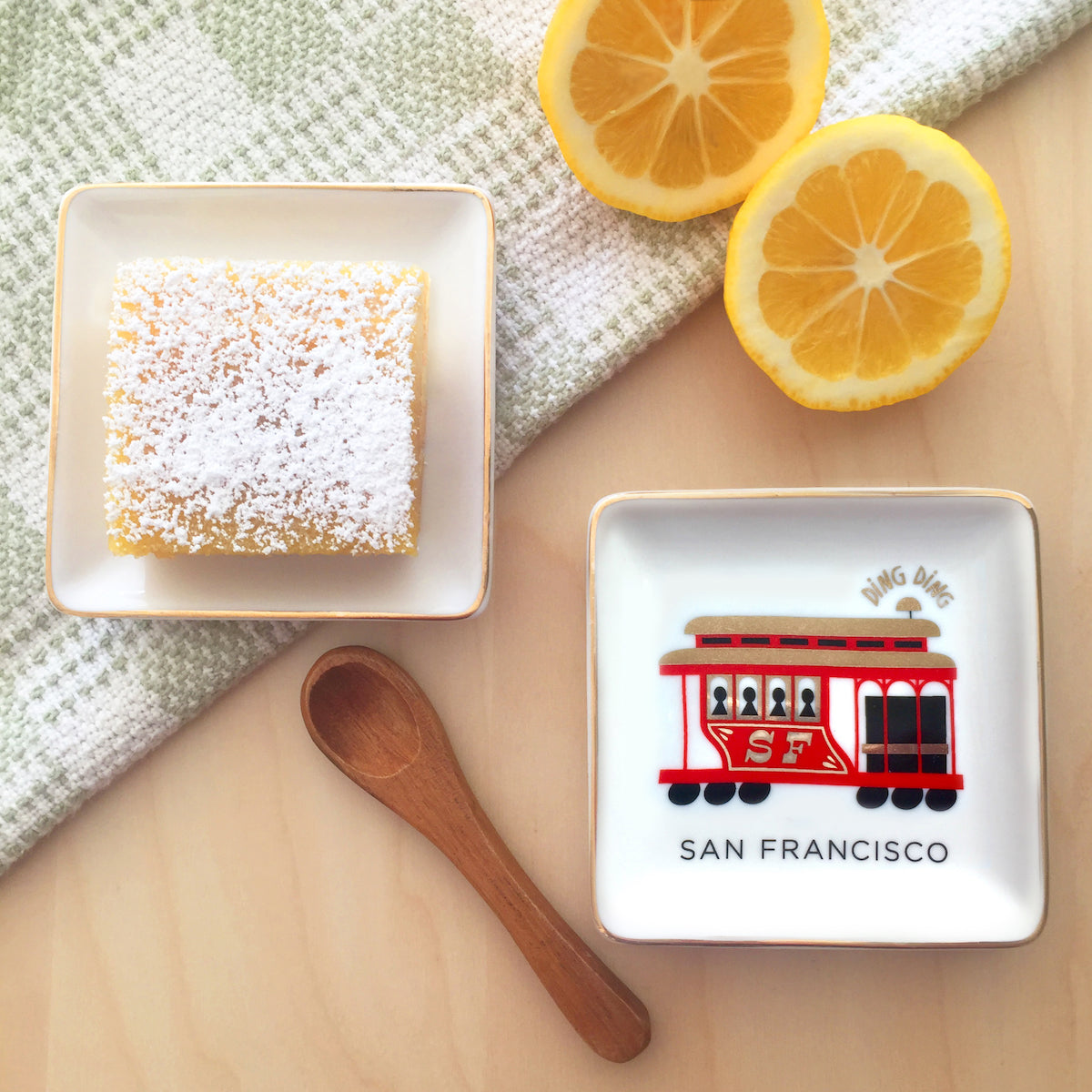 A lemon bar cookie, cut lemons, wooden spoon, tea towel, and small square ceramic dish featuring a colorful illustration of a San Francisco cable car with gold accents sit on a wooden table top.