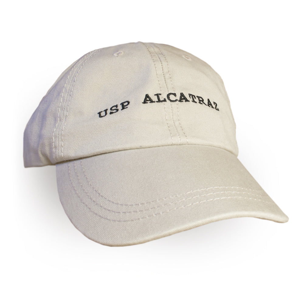 A light tan khaki baseball cap with the words “USP Alcatraz” embroidered on the front panels in black thread.