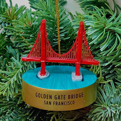 A hand-painted resin Golden Gate Bridge model ornament with gold thread ribbon hangs in front of Christmas foliage.