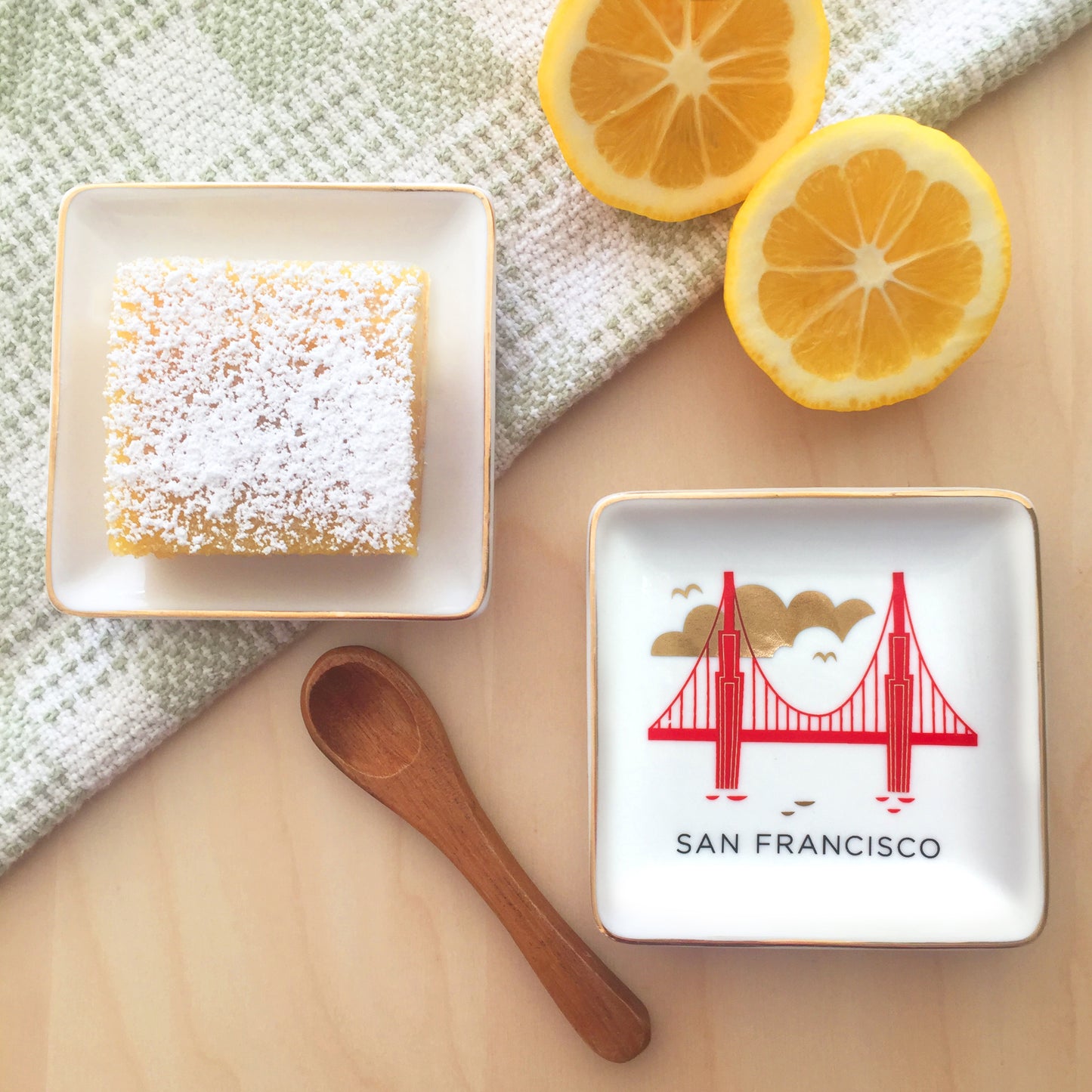 A lemon bar cookie, cut lemons, wooden spoon, tea towel, and small square ceramic dish featuring a colorful illustration of San Francisco’s Golden Gate Bridge with gold accents sit on a wooden table top.