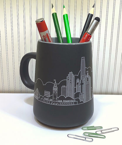 A large grey mug with a San Francisco skyline design is filled with colorful pens and pencils. It sits on a white tabletop next to some paperclips against a striped background.