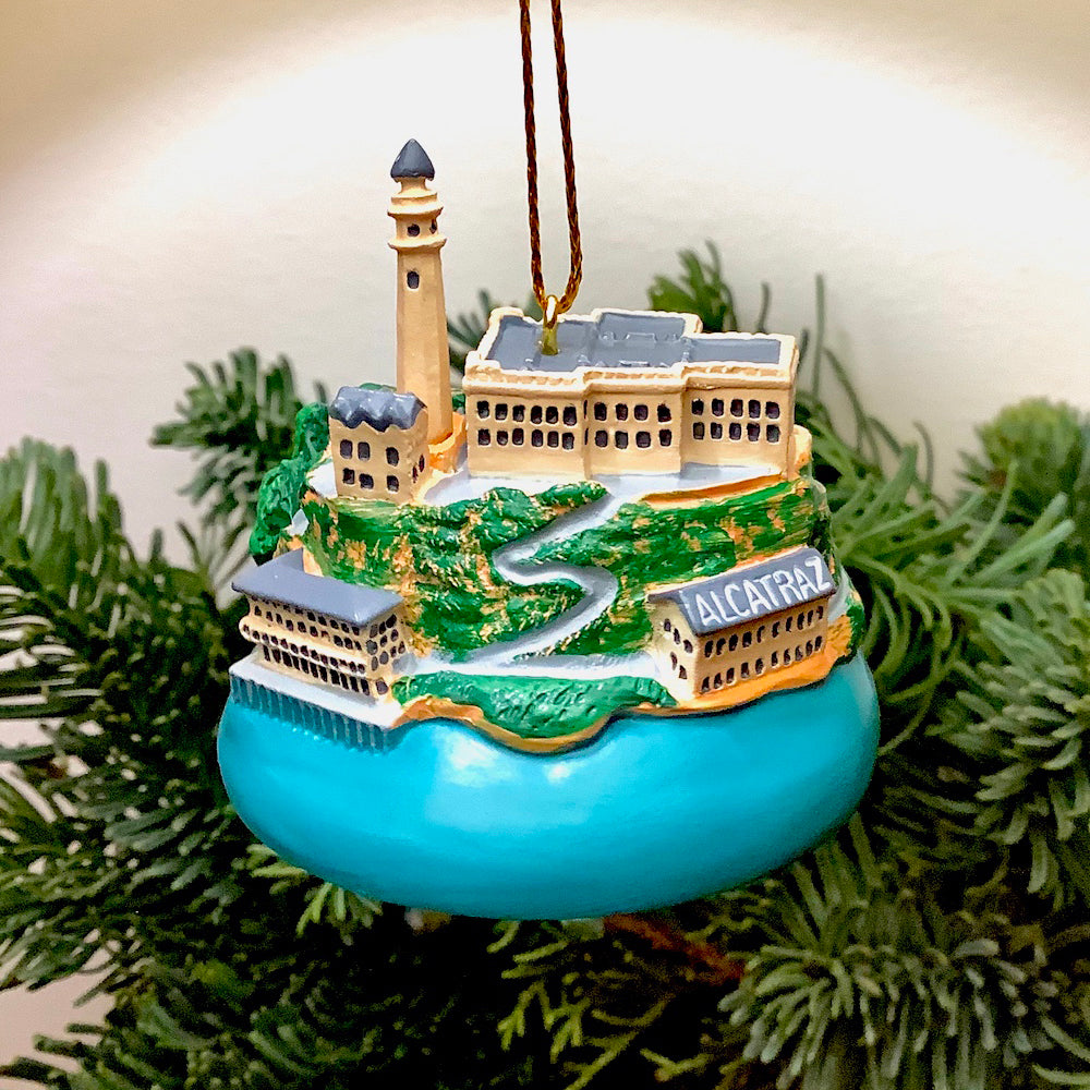 A hand-painted resin Alcatraz Island model ornament with gold thread ribbon hangs in front of Christmas foliage.