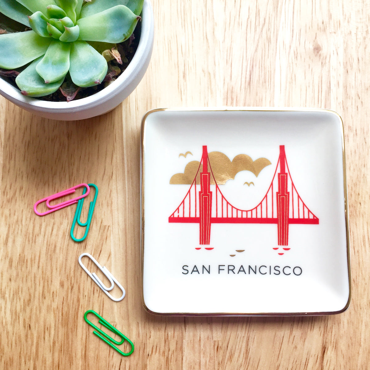 A small succulent, colorful paperclips, and square ceramic dish with a colorful illustration of San Francisco’s Golden Gate Bridge with gold accents sit on a wooden table top.